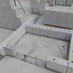 Wznoszenie ścian z białych bloczków betonowych z widocznymi otworami, układanych na zaprawie, z fragmentem ściany z czerwonej cegły w tle.