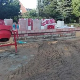 Wznoszenie muru z czerwonych bloczków na fundamencie, widoczna betoniarka i ślady opon na ziemi.