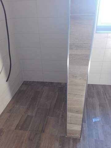 Wykończona kabina prysznicowa z drewnopodobną glazurą na podłodze i ścianach, widoczny odpływ liniowy i aluminiowe profile wykończeniowe.