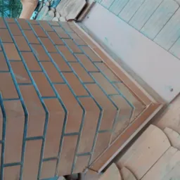 Fragment nowo wybudowanego komina z cegły klinkierowej, połączonego z dachem pokrytym drewnianymi gontami, widoczne metalowe obróbki blacharskie.