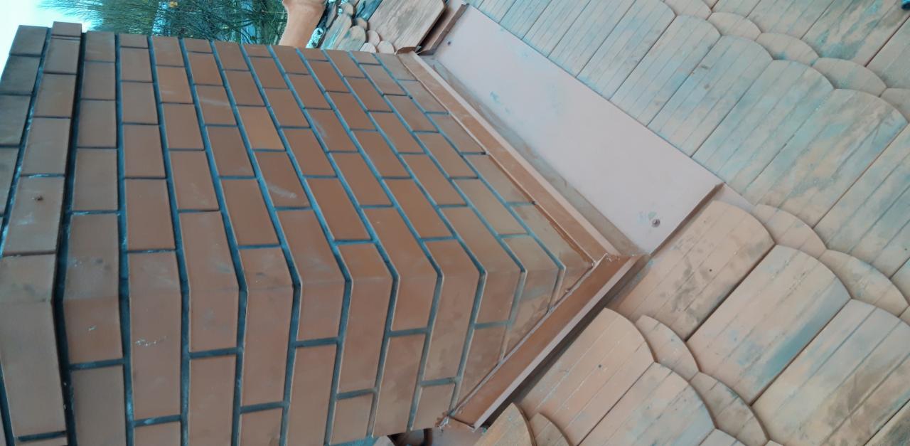 Fragment nowo wybudowanego komina z cegły klinkierowej, połączonego z dachem pokrytym drewnianymi gontami, widoczne metalowe obróbki blacharskie.