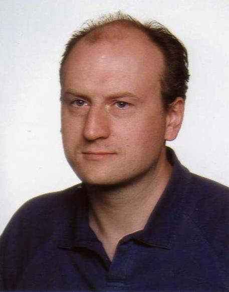 Portret mężczyzny o jasnej karnacji w niebieskiej koszuli polo na jasnym tle. Widoczne zmarszczki mimiczne i zaczesane do tyłu włosy.