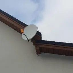 Biała antena satelitarna zamontowana na elewacji domu z brązowym podsufitką, widok z dołu na tle pochmurnego nieba.