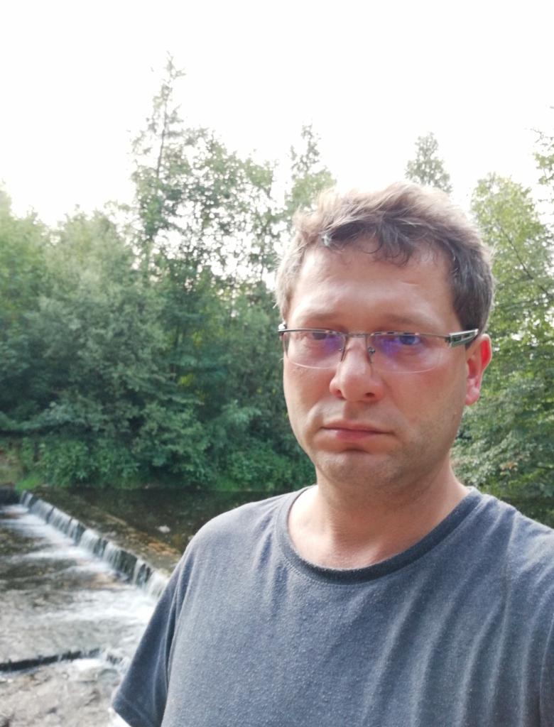 Portret mężczyzny w okularach na tle zieleni i małego wodospadu, z widocznym fragmentem szarej koszulki. Zdjęcie z bliska, naturalne światło.