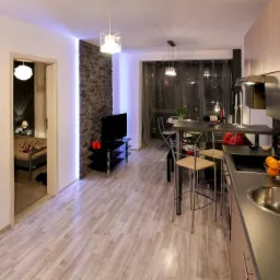 Nowoczesne wnętrze z drewnopodobną podłogą, kamienną ścianą z podświetleniem LED i aneksem kuchennym z szarym blatem oraz owocami w misce.