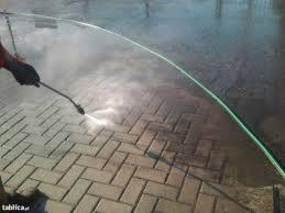 Czyszczenie kostki brukowej myjką ciśnieniową, widoczny strumień wody usuwający zabrudzenia, fragment węża i ręka operatora.