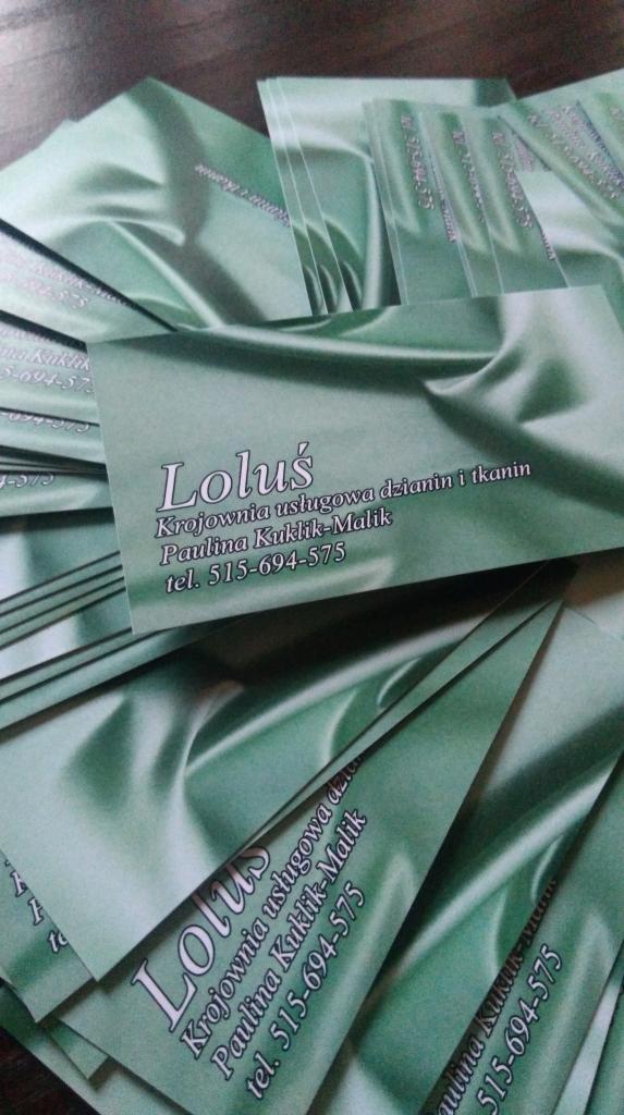 Stos wizytówek firmy krawieckiej Lolus na tle materiału w odcieniach zieleni, z widocznym logo i danymi kontaktowymi.