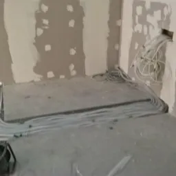 Wnętrze w trakcie prac wykończeniowych, widoczne peszle elektryczne ułożone wzdłuż podłogi i wychodzące ze ściany, ściany częściowo wykończone gładzią gipsową, zwój kabli przy ścianie, widoczny...