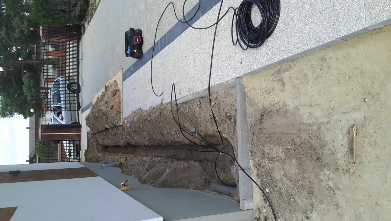 Wykop w ziemi z ułożonymi przewodami elektrycznymi, widoczna rolka kabla, torba narzędziowa i fragment posesji z ogrodzeniem.