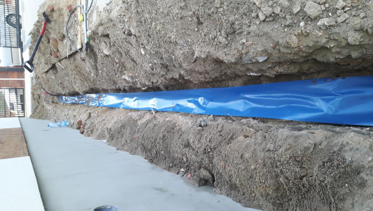 Wykop w ziemi z niebieską folią ochronną, prawdopodobnie zabezpieczającą kable elektryczne, widoczne przewody i narzędzia w tle.
