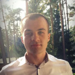 Młody mężczyzna w białej koszuli z wzorem w kratę na kołnierzyku, na tle iglastego lasu w słoneczny dzień.