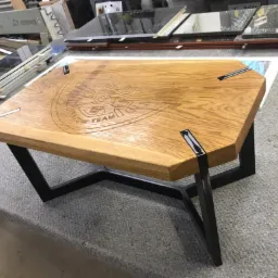 Stolik kawowy z blatem z jasnego drewna z wygrawerowanym logo 'UHD Kraków Team', osadzony na czarnych, metalowych nogach o geometrycznym kształcie.