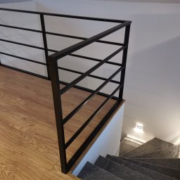 HolMat - Nowoczesna, czarna, metalowa barierka z poziomymi szczeblami zabezpieczająca schody wewnątrz domu, widok z góry na drewnianą podłogę i schody pokryte szarą wykładziną.