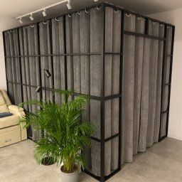 HolMat - Metalowa konstrukcja z szarymi zasłonami tworząca ściankę działową w nowoczesnym wnętrzu, oświetlona reflektorami sufitowymi, z rośliną doniczkową w szarej osłonce na pierwszym planie.