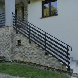 HolMat - Zewnętrzne schody z szarego kamienia prowadzące do domu, z nowoczesną, poziomą balustradą z ciemnego metalu, otoczone zielonym trawnikiem i rabatami kwiatowymi.