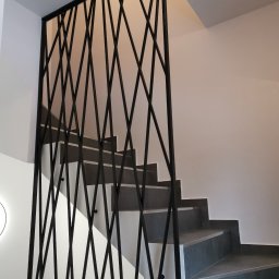 HolMat - Nowoczesne schody z szarymi stopniami i geometryczną, czarną balustradą z metalowych prętów na tle białych ścian.