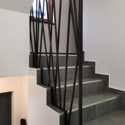 HolMat - Nowoczesne schody z szarymi płytkami i czarną, geometryczną balustradą, widok z dołu na górę, z butami na kaloryferze w tle.