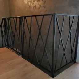 Czarna poręcz metalowa z geometrycznym wzorem krzyżujących się prętów na tle ściany z efektem dekoracyjnym i drewnianej podłogi.