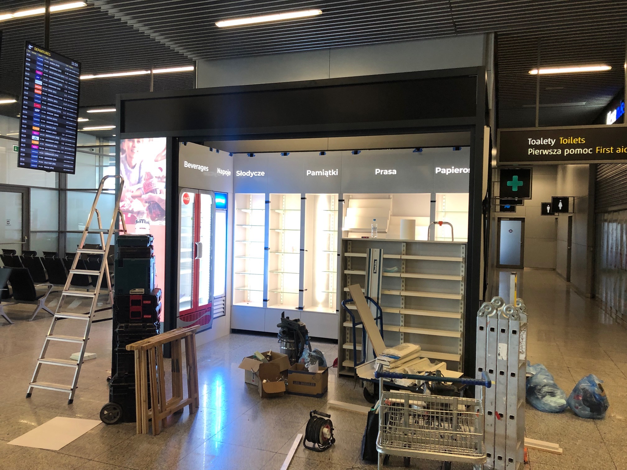 Montaż lub remont stoiska handlowego na lotnisku, widoczne regały z podświetleniem, drabiny i narzędzia, w tle tablica odlotów.
