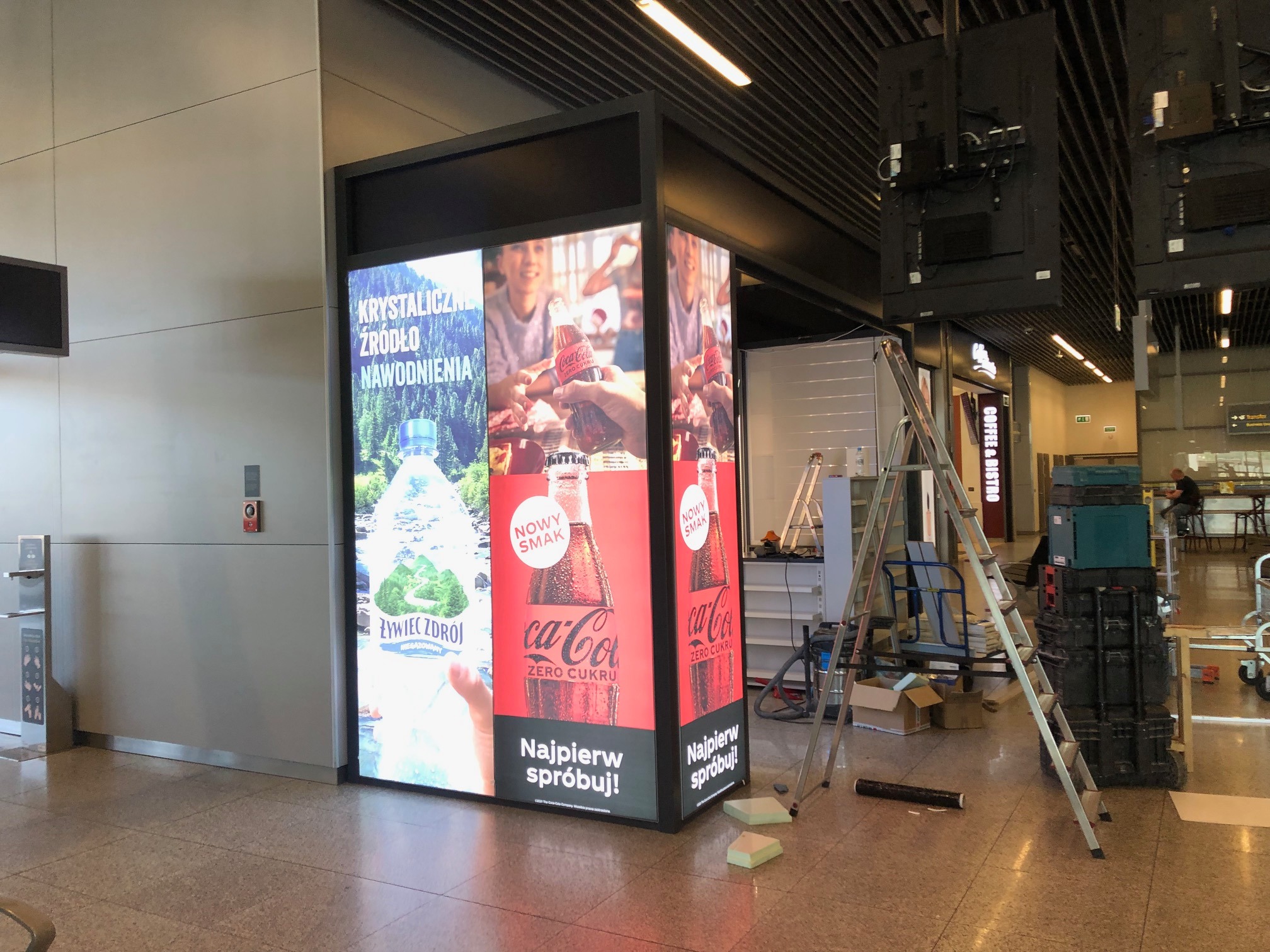 Montaż podświetlanej reklamy z napojami w przestrzeni publicznej, widoczna drabina, skrzynki narzędziowe i elementy konstrukcyjne.