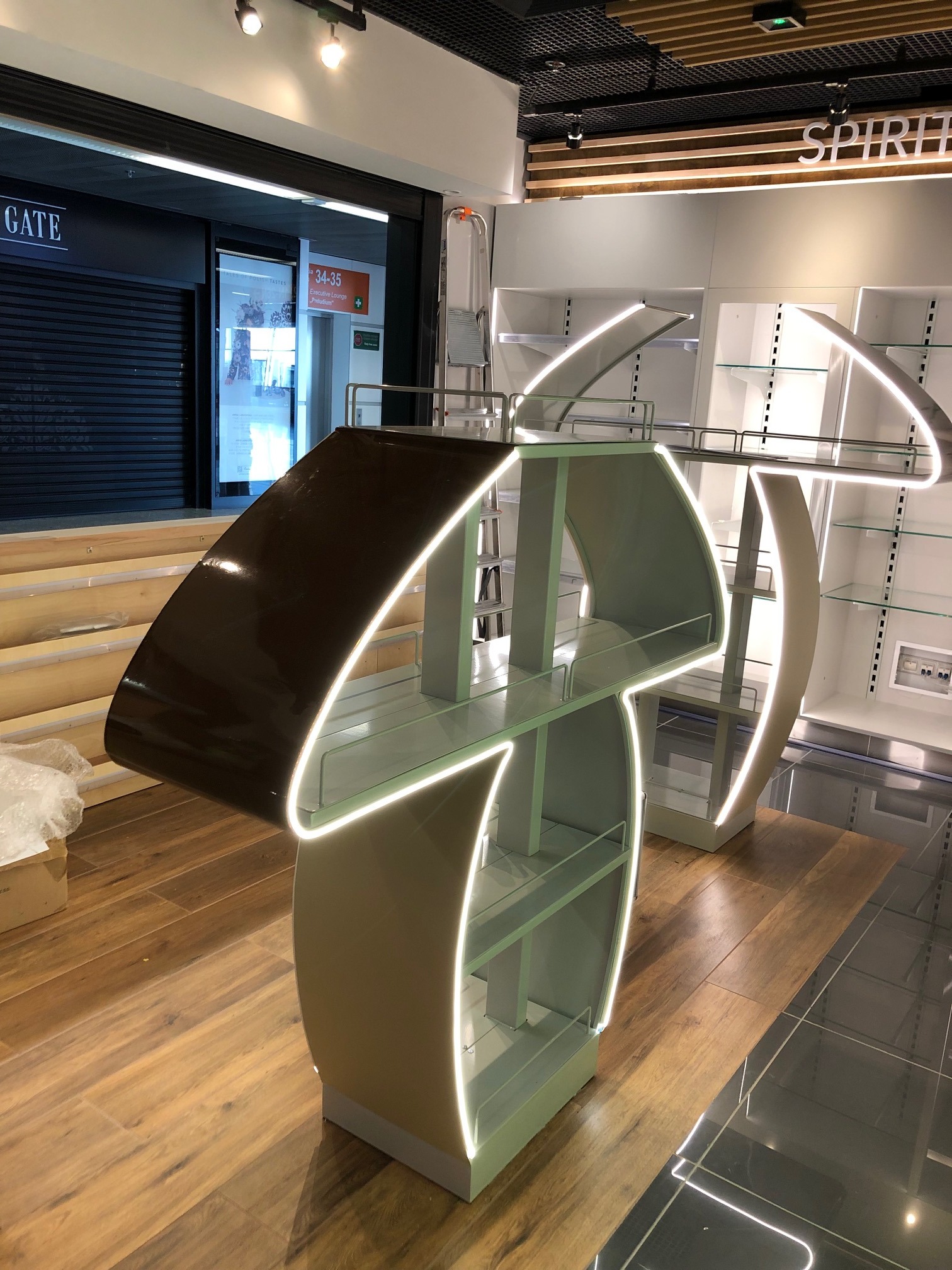 Nowoczesna, podświetlana konstrukcja metalowa z półkami szklanymi w sklepie, prawdopodobnie element wyposażenia stoiska handlowego, z widocznymi elementami metalowymi i oświetleniem LED.