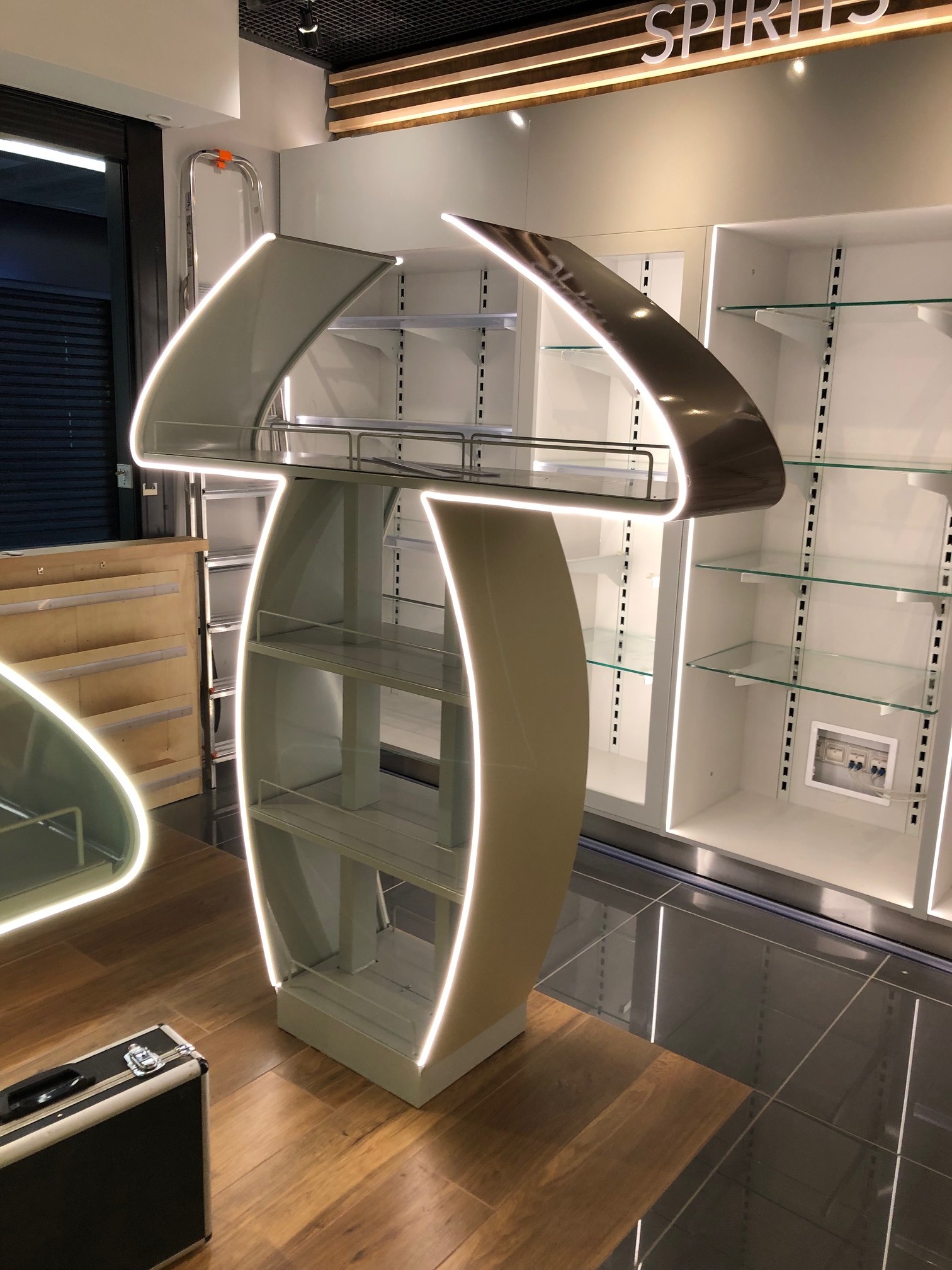 Metalowa konstrukcja ekspozycyjna z podświetleniem LED, półki szklane, tło stanowią białe regały sklepowe, na podłodze ciemne płytki.