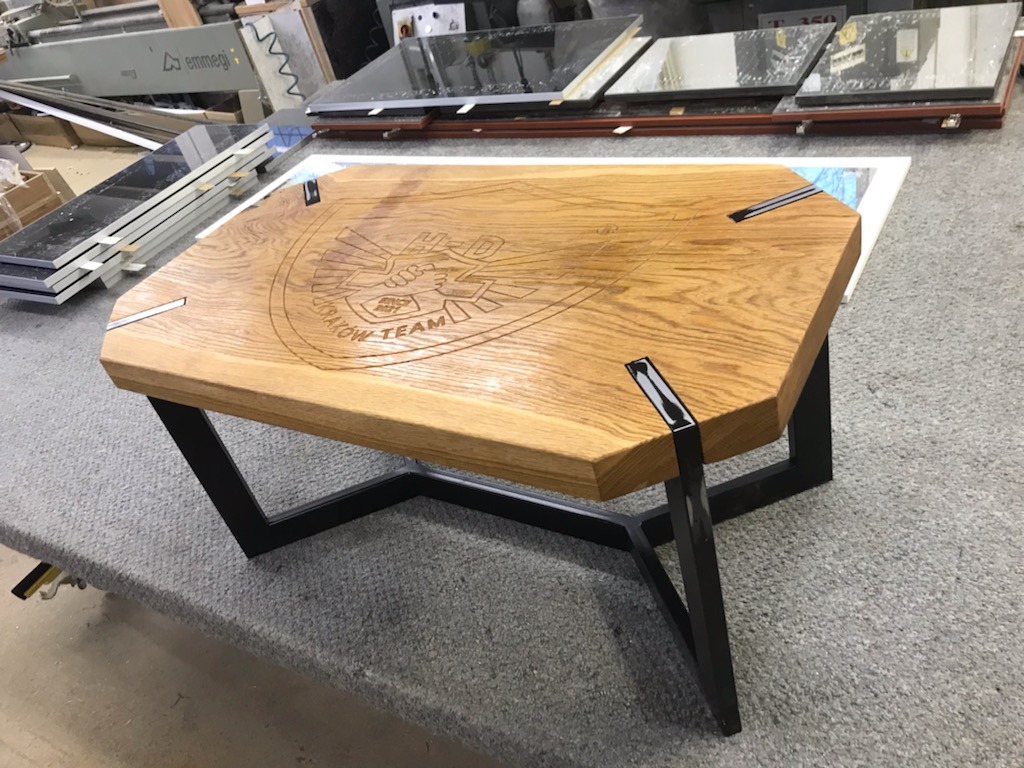 Stolik kawowy z blatem z jasnego drewna z wygrawerowanym logo 'UHD Kraków Team', osadzony na czarnych, metalowych nogach o geometrycznym kształcie.