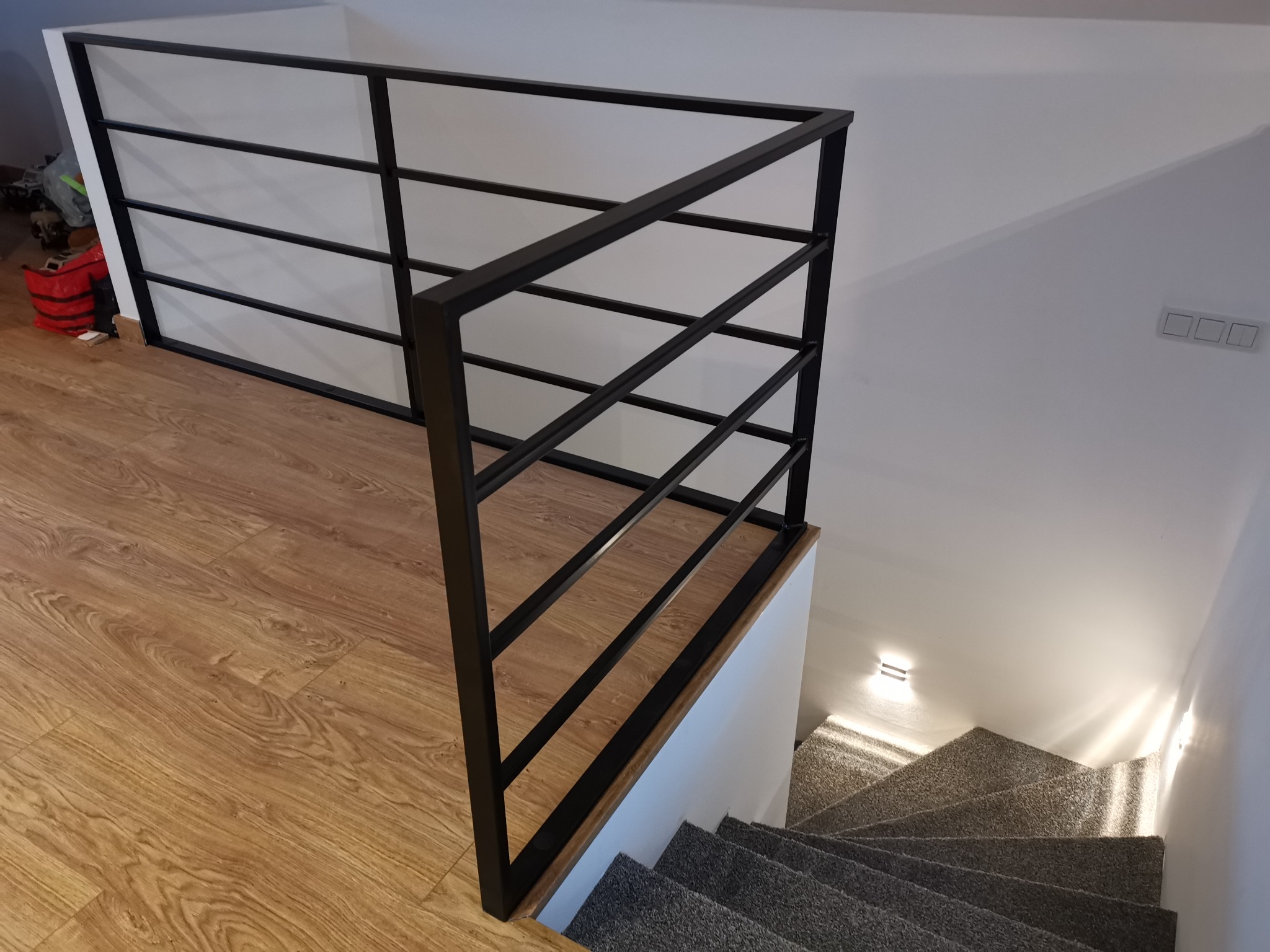 Nowoczesna, czarna, metalowa barierka z poziomymi szczeblami zabezpieczająca schody wewnątrz domu, widok z góry na drewnianą podłogę i schody pokryte szarą wykładziną.