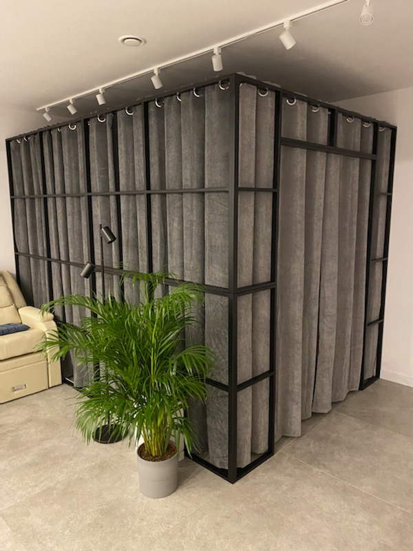 Metalowa konstrukcja z szarymi zasłonami tworząca ściankę działową w nowoczesnym wnętrzu, oświetlona reflektorami sufitowymi, z rośliną doniczkową w szarej osłonce na pierwszym planie.