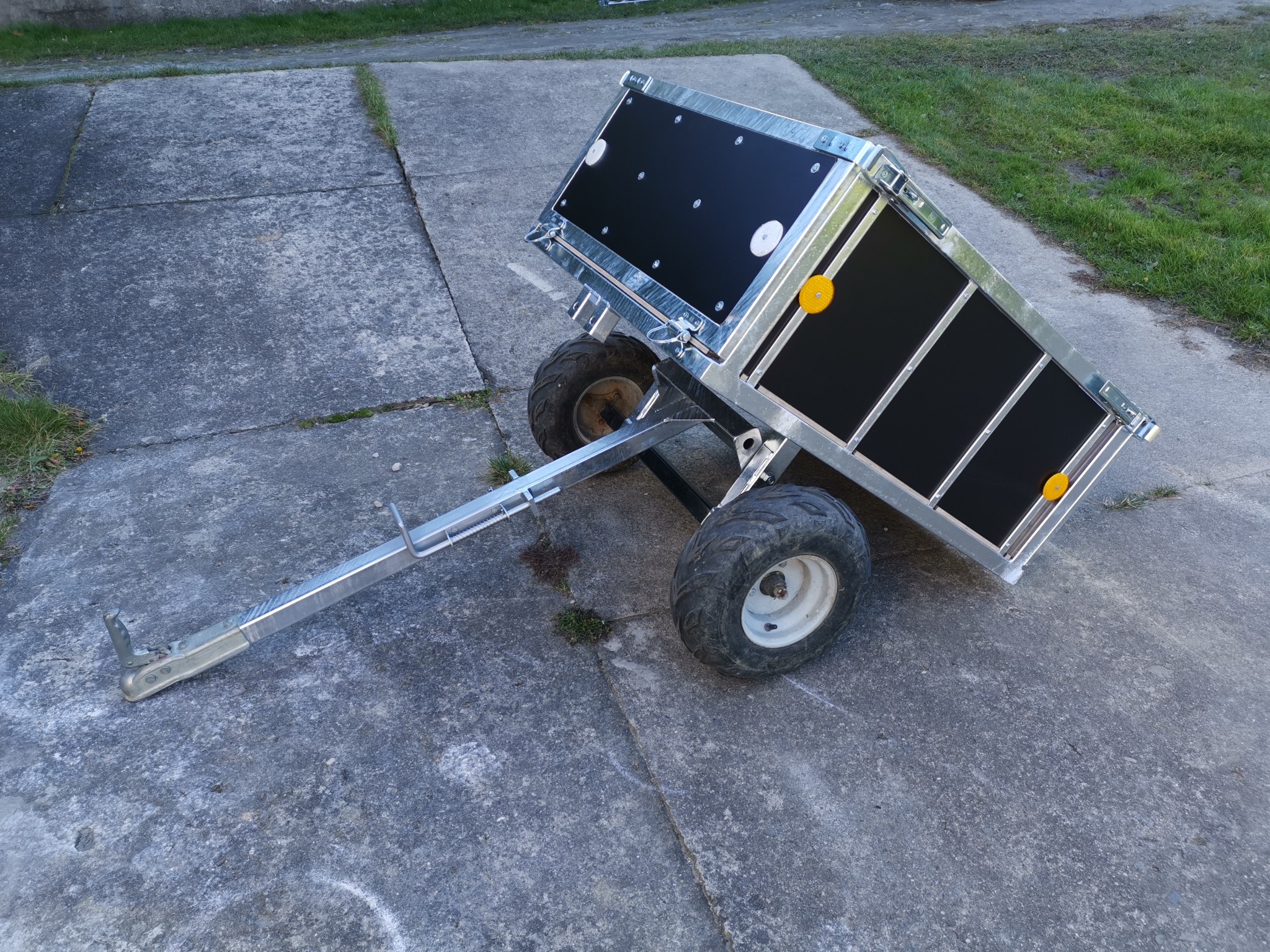 Nowa, metalowa przyczepka z czarnymi panelami, stojąca na betonowej nawierzchni, z widocznymi oponami terenowymi i elementami mocującymi.