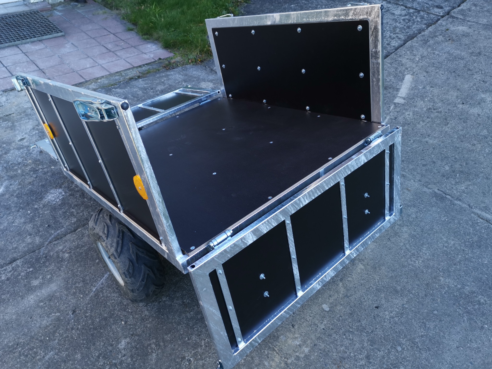 Przyczepka metalowa z czarnym wykończeniem, widoczne koła terenowe i srebrne okucia, stojąca na betonowym podłożu.