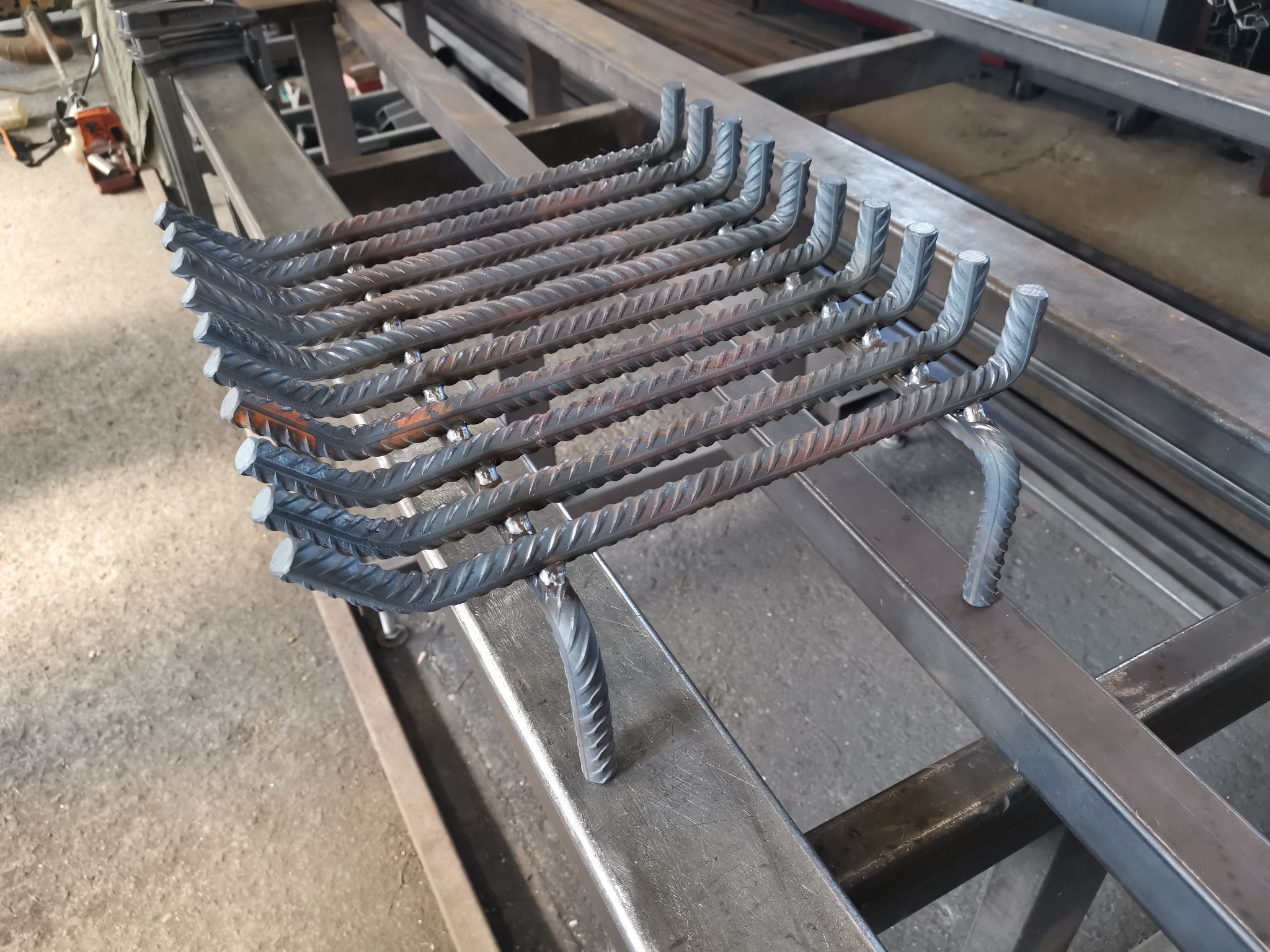 Konstrukcja z prętów żebrowanych, spawana w kształcie drabinki z wygiętymi końcami, leżąca na metalowym stelażu w warsztacie.