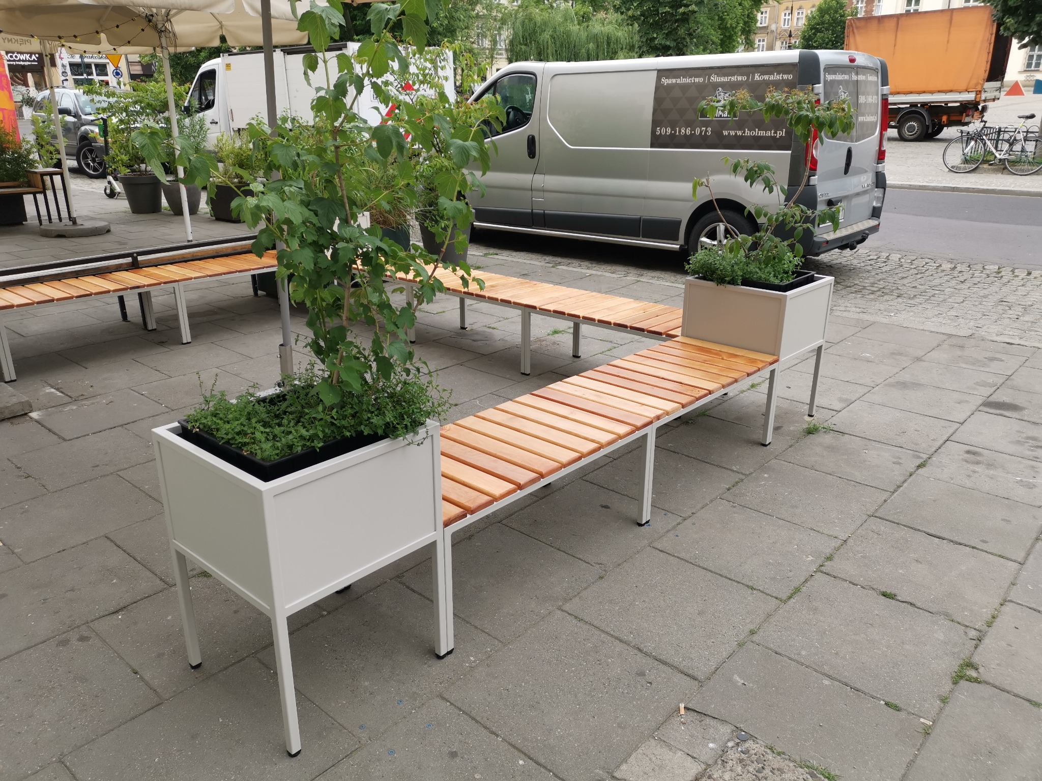 Ławka miejska z drewnianym siedziskiem i wbudowanymi donicami z zielenią, zrealizowana w Krzywaczce. W tle zaparkowany bus z reklamą firmy.
