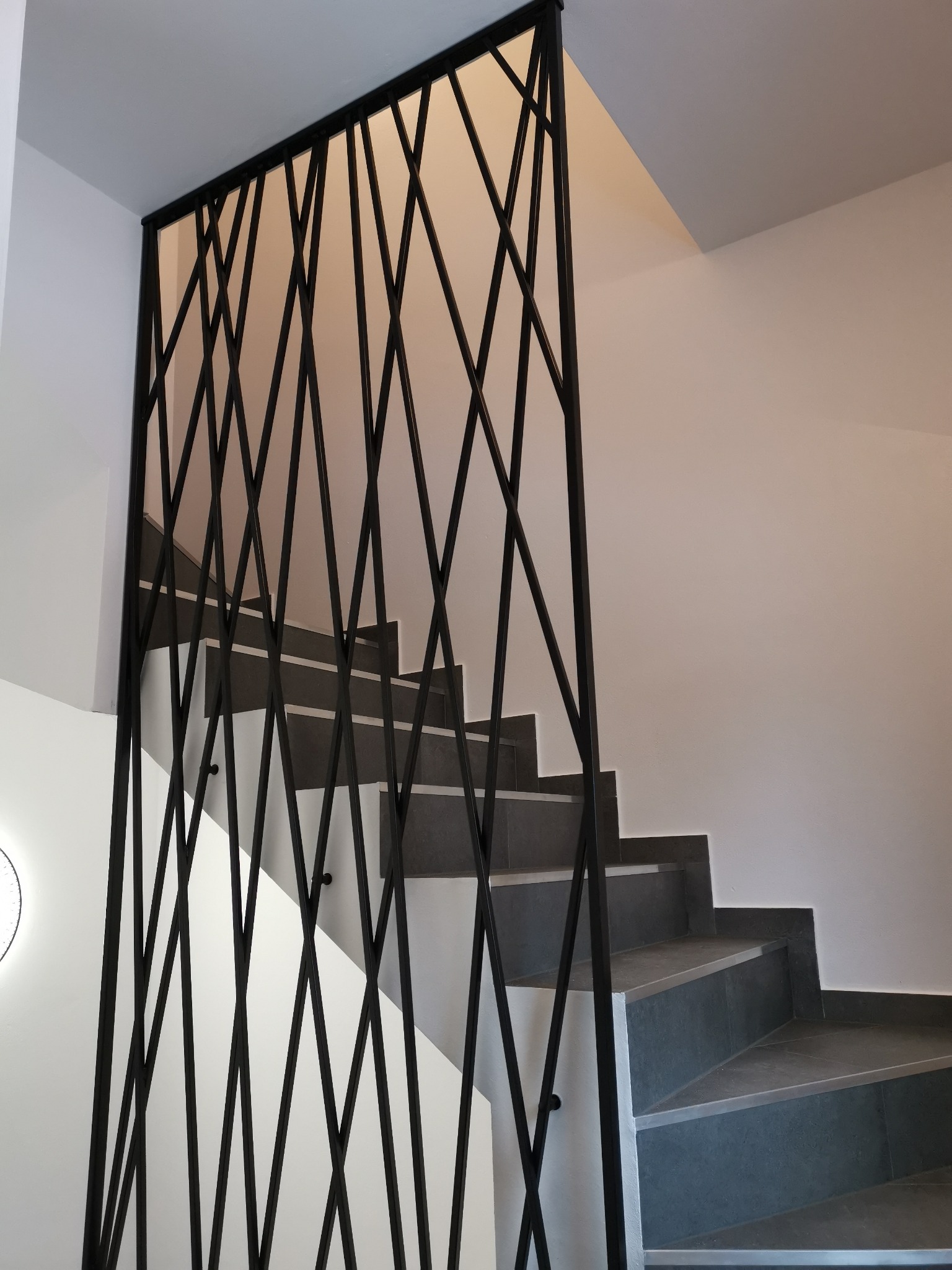 Nowoczesne schody z szarymi stopniami i geometryczną, czarną balustradą z metalowych prętów na tle białych ścian.