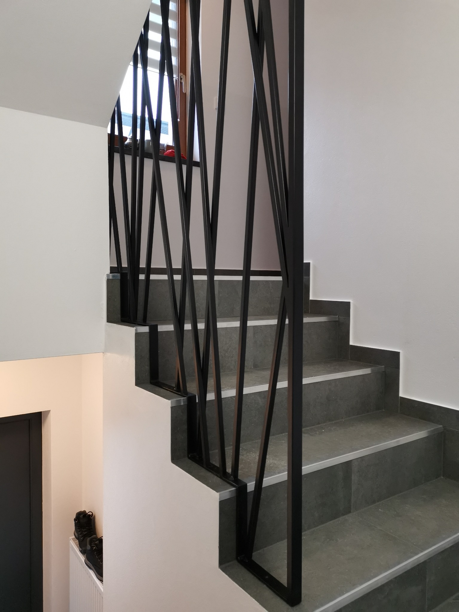 Nowoczesne schody z szarymi płytkami i czarną, geometryczną balustradą, widok z dołu na górę, z butami na kaloryferze w tle.
