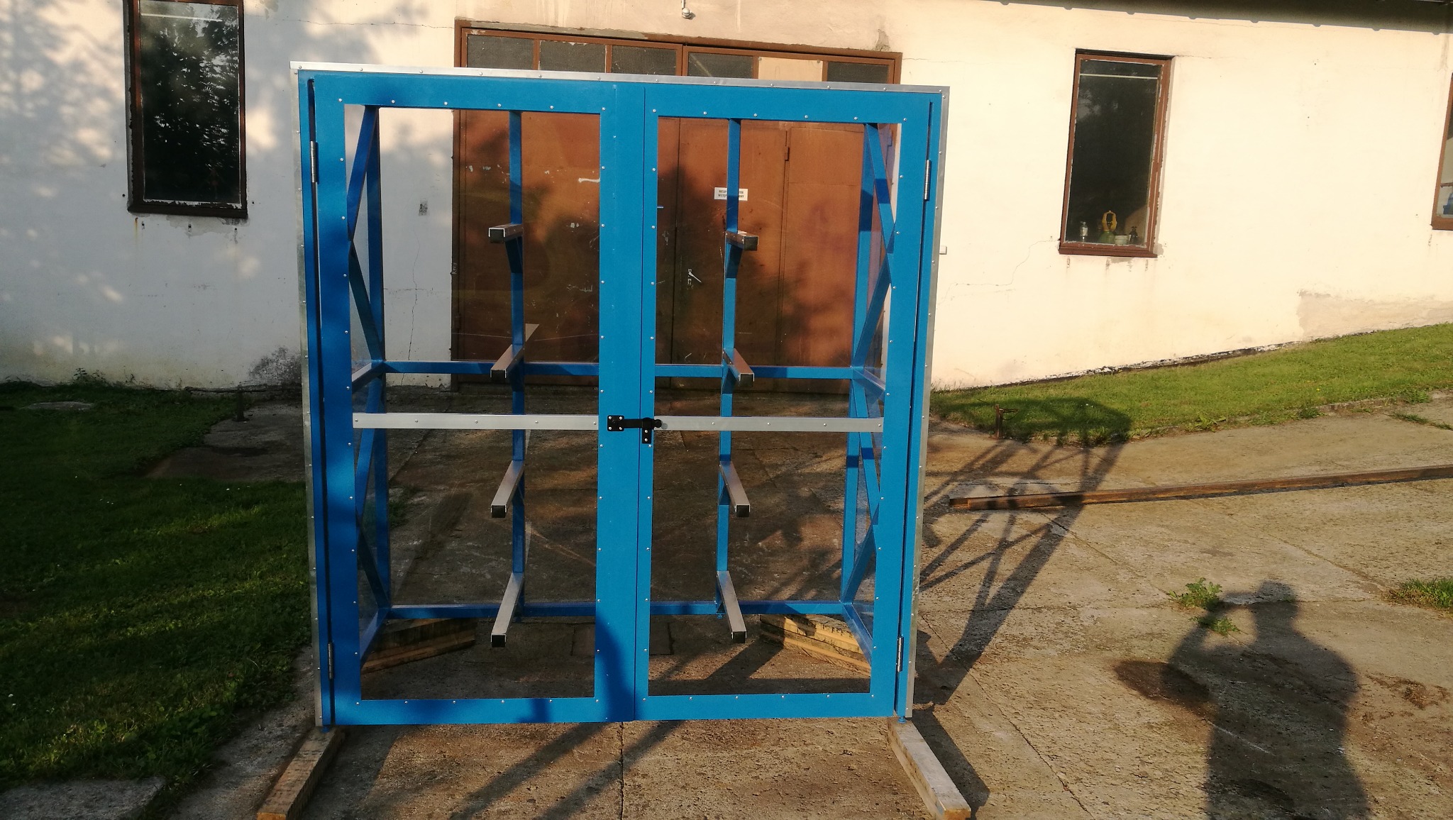Niebieska metalowa konstrukcja z zamkiem i srebrnymi poprzeczkami, ustawiona na drewnianych belkach na betonowym podłożu przed białym budynkiem z brązowymi drzwiami i oknami, cień fotografa...