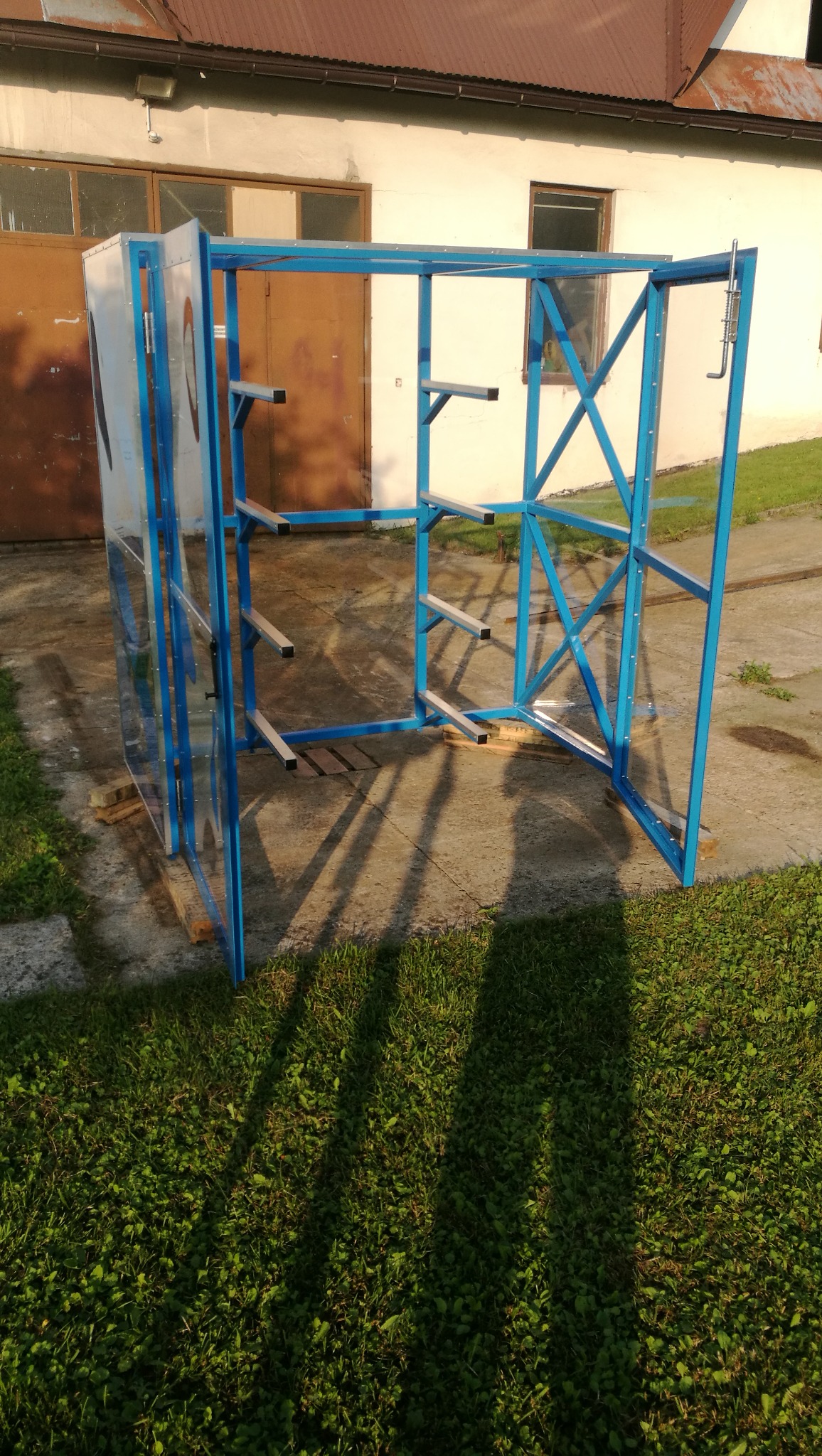 Niebieska metalowa konstrukcja z półkami i przezroczystym panelem bocznym, stojąca na trawie przed budynkiem.