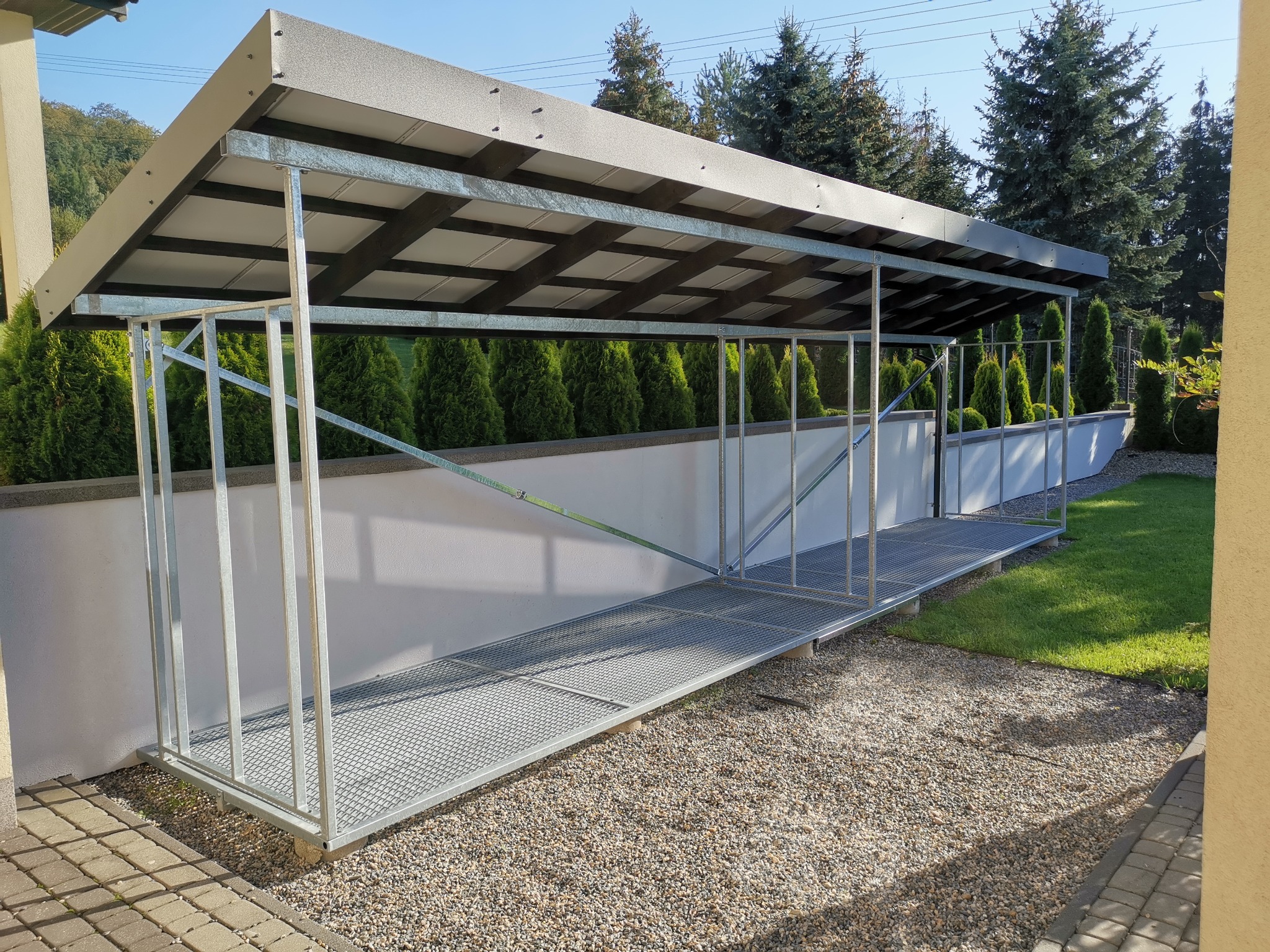 Metalowa konstrukcja z zadaszeniem, prawdopodobnie wiata lub zadaszone przejście, wykonana ze stalowych profili i kratownicy, umieszczona na żwirowym podłożu obok białego murku i zielonego trawnika.