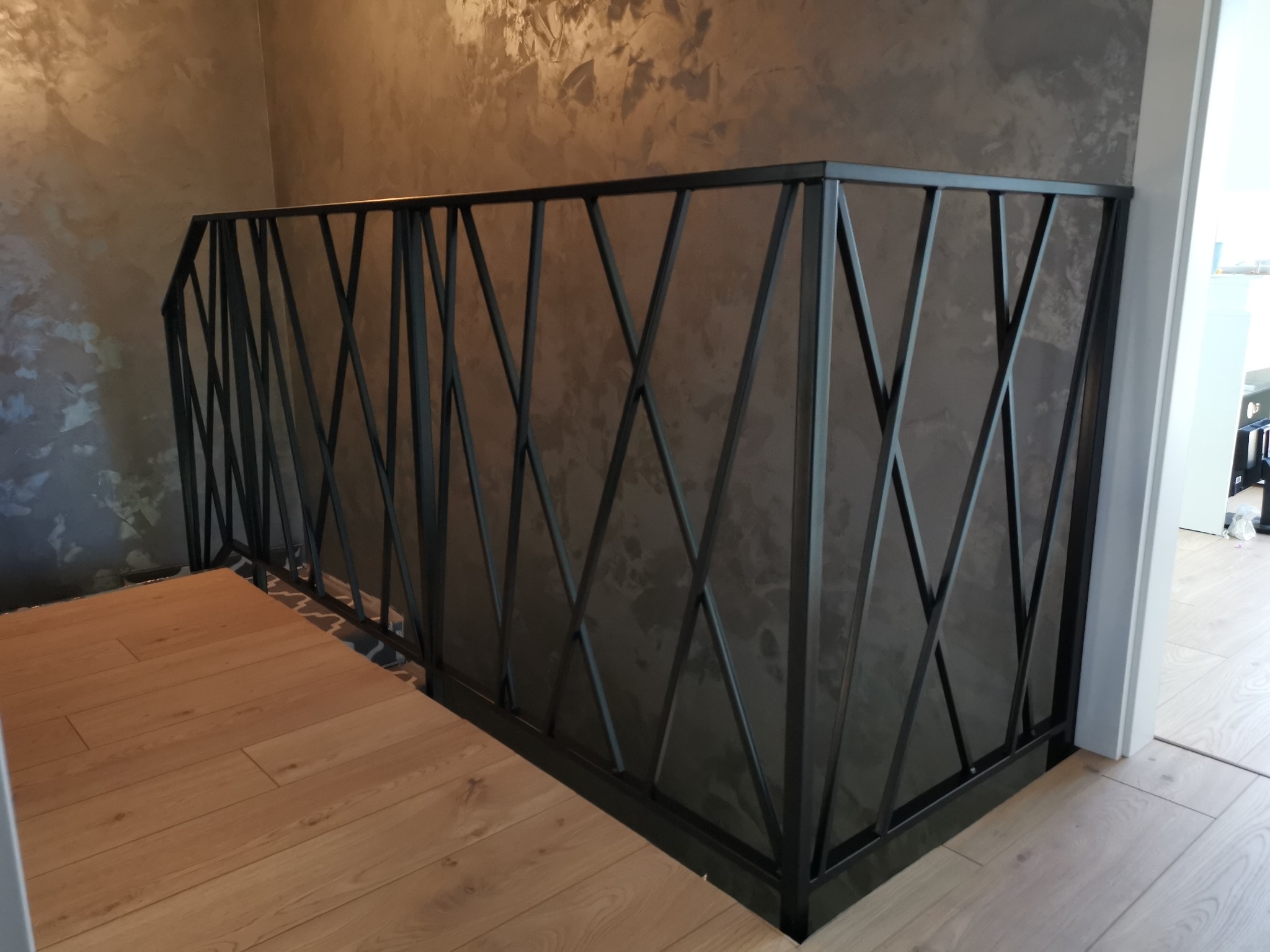 Czarna poręcz metalowa z geometrycznym wzorem krzyżujących się prętów na tle ściany z efektem dekoracyjnym i drewnianej podłogi.