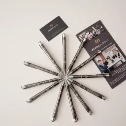 Układ pióra, wizytówki i ulotki firmy Infinite Business na białym tle. Pióra ułożone promieniście wokół centralnego punktu. Wizytówka i ulotka w kolorze ciemnoszarym z białym tekstem i logo firmy.