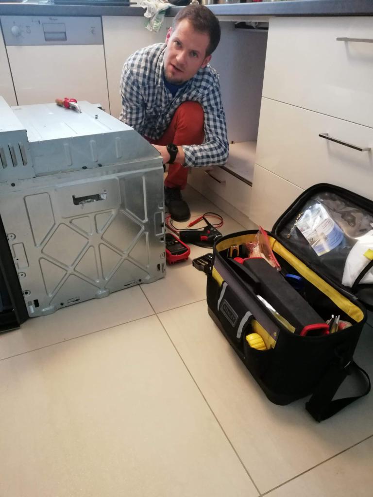 Mężczyzna w kraciastej koszuli i czerwonych spodniach kuca obok wyjętego piekarnika, z otwartą skrzynką narzędziową Stanley obok, w tle białe szafki kuchenne i zmywarka.