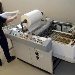 Pracownik drukarni obsługuje maszynę drukarską Delta, z rolą papieru i stosem wydrukowanych arkuszy z powtarzalnym wzorem.