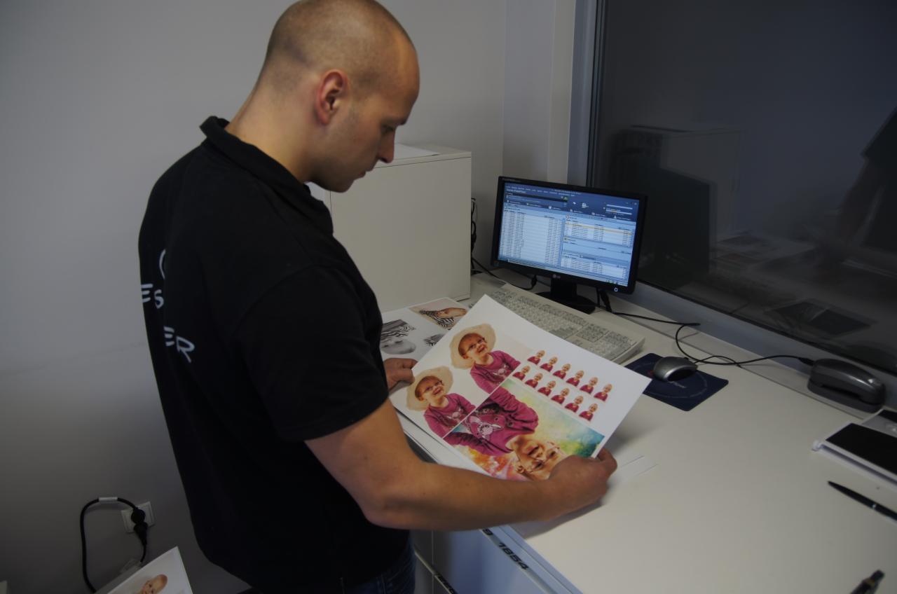Pracownik drukarni przegląda arkusz z wydrukowanymi zdjęciami dziecka w różnych ujęciach, na biurku komputer i inne arkusze z grafikami.
