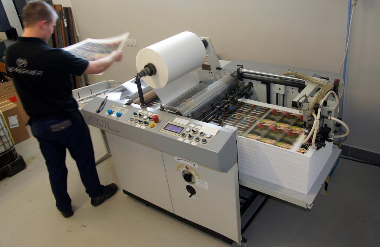 Pracownik drukarni obsługuje maszynę drukarską Delta, z rolą papieru i stosem wydrukowanych arkuszy z powtarzalnym wzorem.