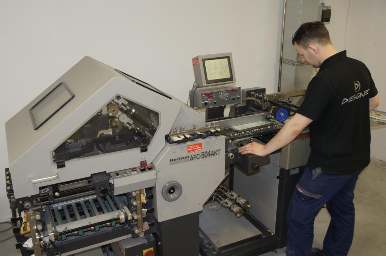 Pracownik obsługuje maszynę Horizon AFC-504AKT do składania arkuszy w drukarni, widoczny ekran sterujący i mechanizm podawania papieru.