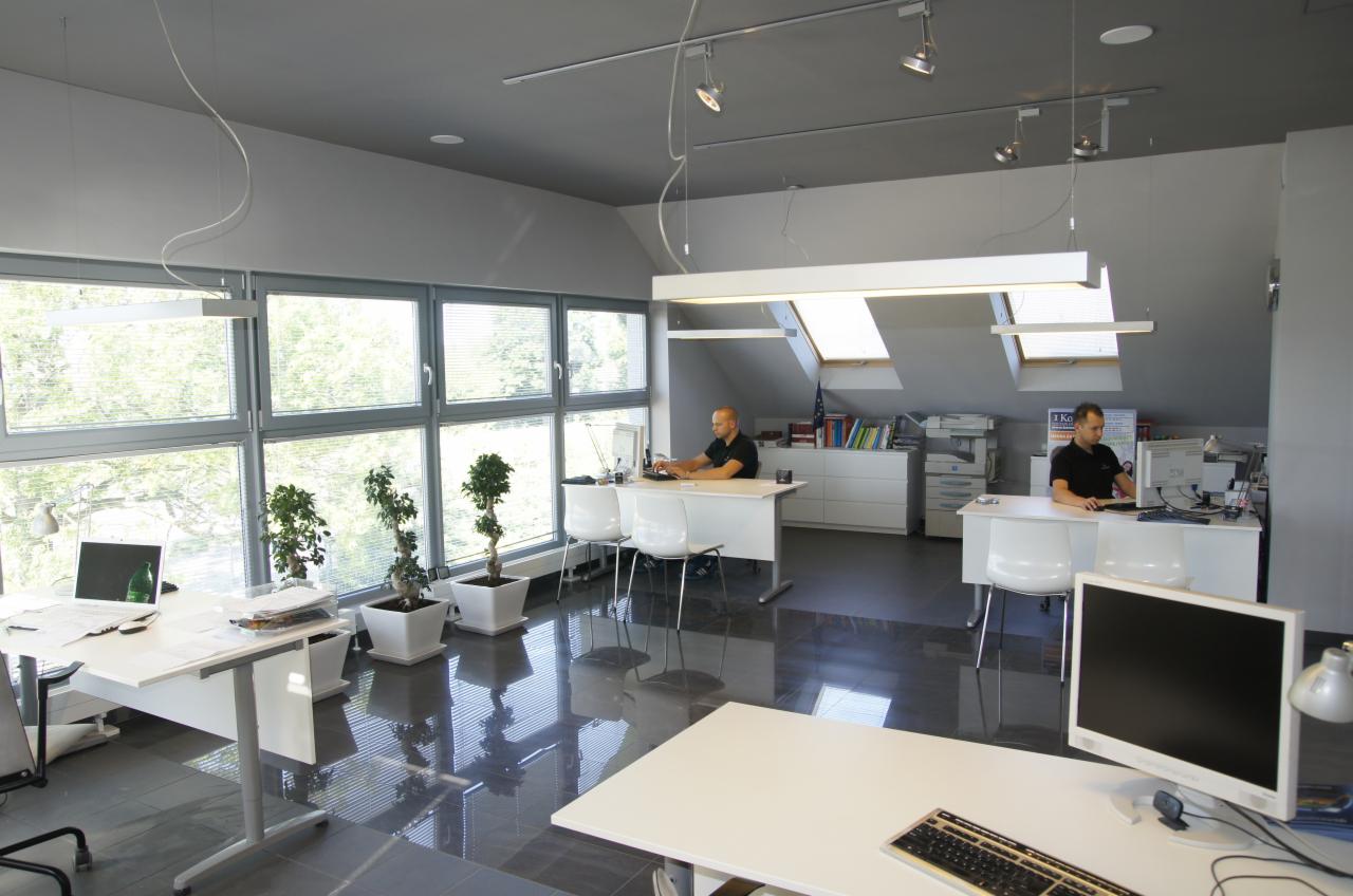 Przestronne biuro na poddaszu z dużymi oknami, białymi biurkami i pracownikami przy komputerach, widoczne rośliny doniczkowe i nowoczesne oświetlenie.