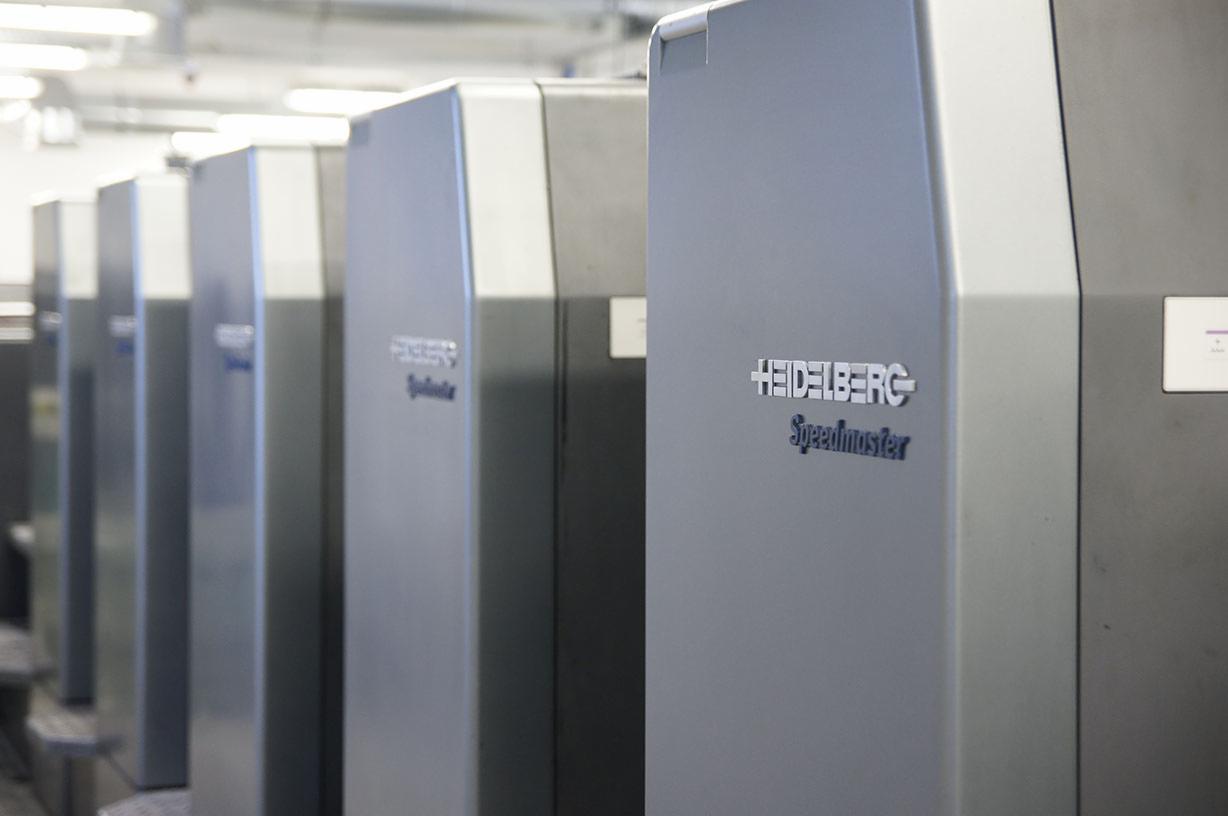 Rząd szarych maszyn drukarskich Heidelberg Speedmaster w hali produkcyjnej, widok z boku, delikatne oświetlenie.