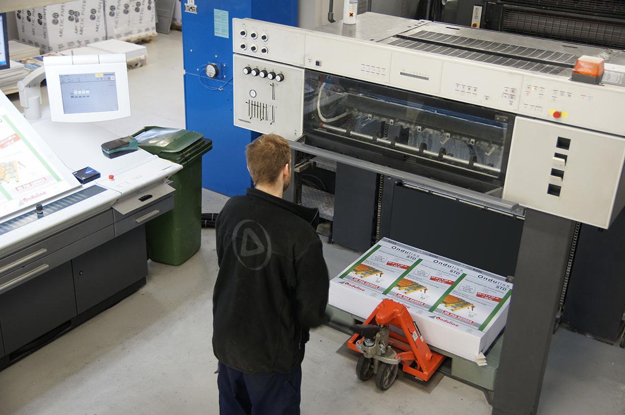 Operator w czarnej bluzie z logo firmy nadzoruje proces druku arkuszy z grafiką na dużej maszynie drukarskiej, stos arkuszy na wózku paletowym.