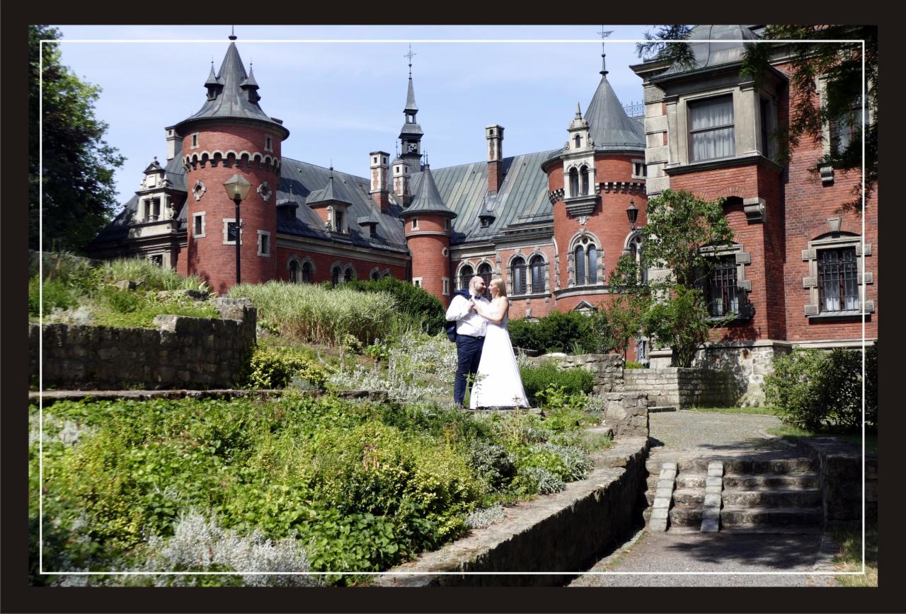 Para młoda pozuje na tle ceglanego zamku z wieżami i zielonym ogrodem.