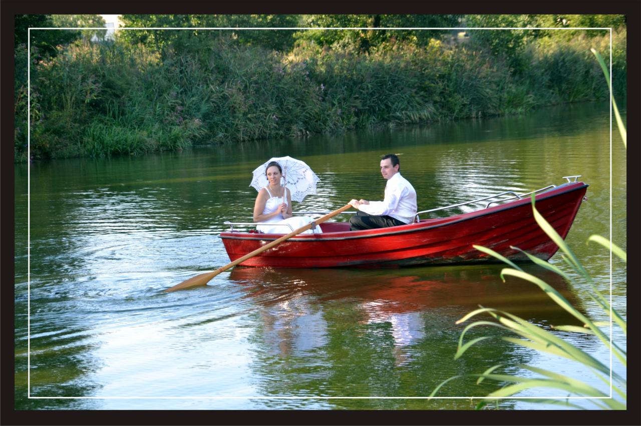 Para młoda płynie łódką po jeziorze; panna młoda trzyma biały parasol, pan młody wiosłuje.